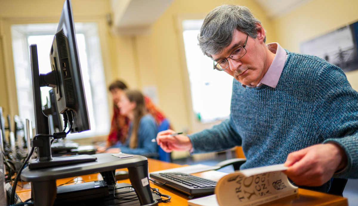 A customer doing some research about his Scottish heritage. He is sitting at a desk, in front of a computer and also looking at a book.