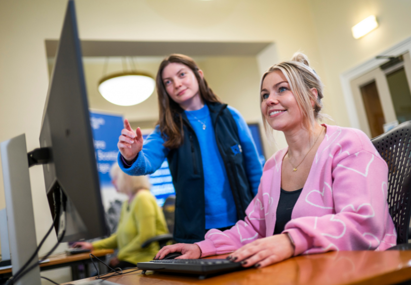 Woman smiles looking at a computer screen while another woman points at the screen
