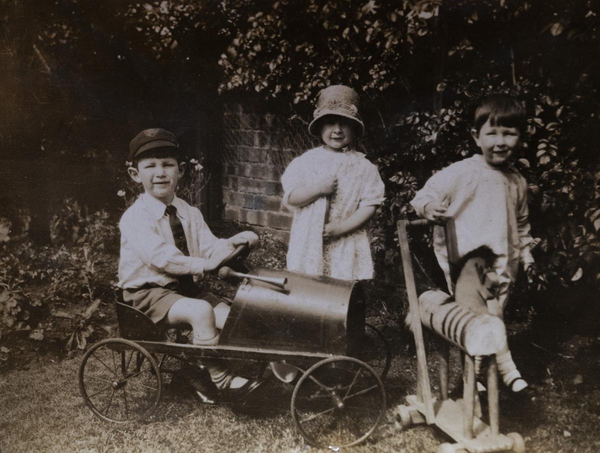 3 children playing in a garden standing in front of a brick wall. 1 is in a toy car, another has a wooden horse.