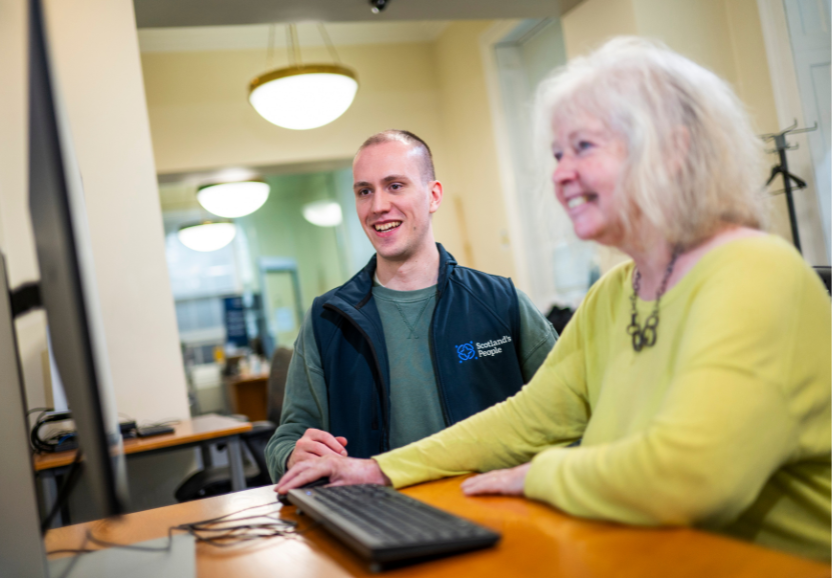 A woman and man look at a computer smiling, seated at a desk
